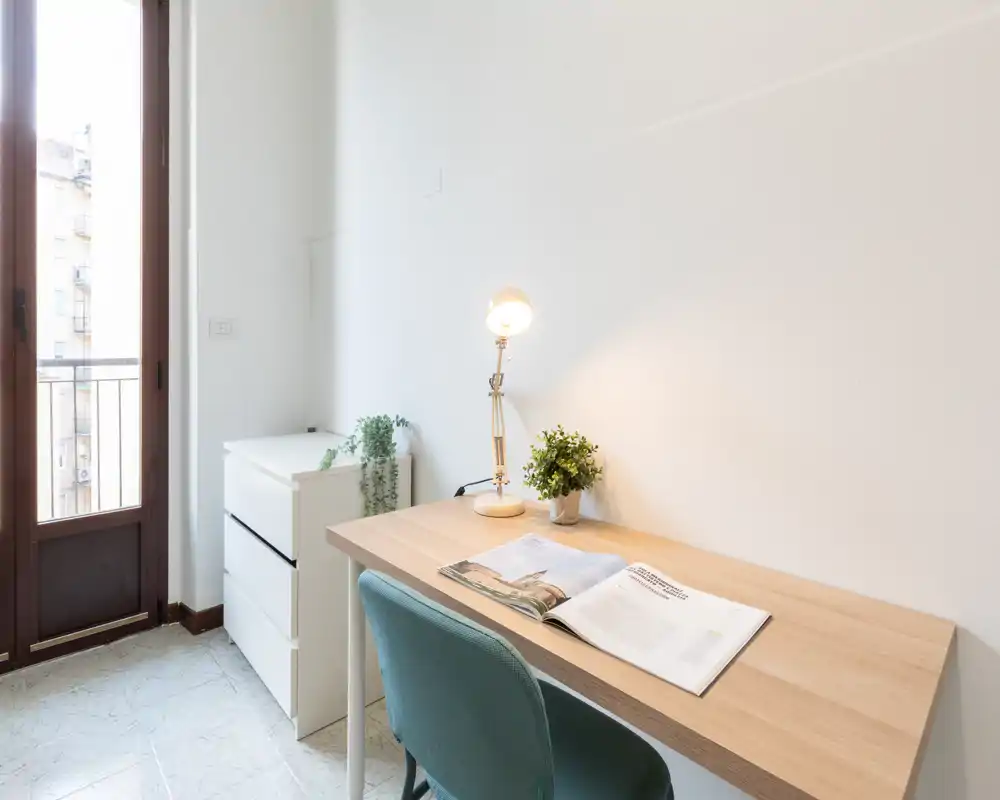 Coin bureau lumineux près d'une grande fenêtre avec un bureau en bois, une chaise turquoise, une lampe et de petites plantes — un espace calme idéal pour étudier ou télétravailler.