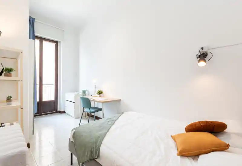 Chambre lumineuse et minimaliste avec lit bien fait, coussins chaleureux et petit bureau près de la porte du balcon — parfaite pour un séjour confortable.