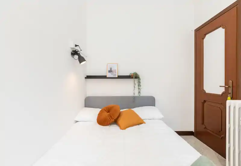 Piccola camera da letto luminosa con un letto matrimoniale ben rifatto, cuscini dai toni caldi e una mensola minimal con pianta decorativa. Pareti bianche e luce naturale per un'atmosfera tranquilla e accogliente.