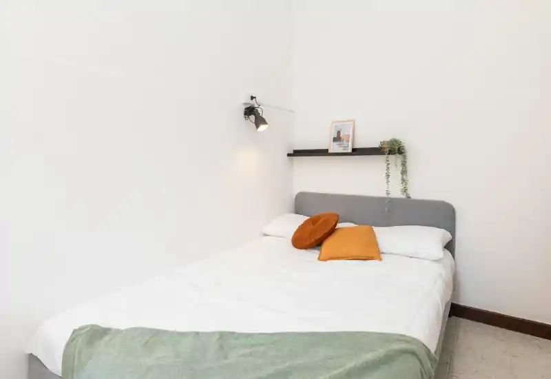Chambre lumineuse et minimaliste avec un lit double en linge blanc, deux coussins décoratifs et un plaid ; une lampe murale et une étagère avec plante apportent une touche moderne.