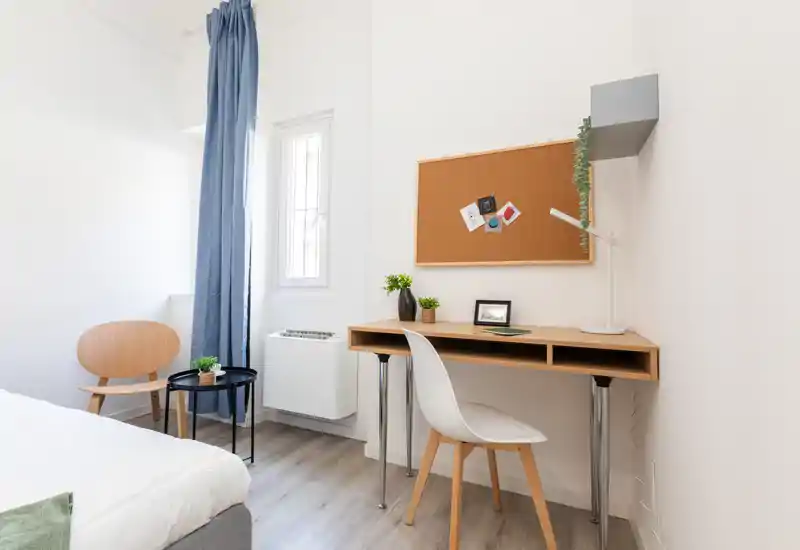 Bright, modern bedroom corner with a neatly made bed, a small wooden desk and a comfortable chair by a window. Minimal, airy styling with plants and a cork noticeboard adds a cozy yet functional study area.