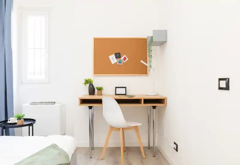 Bright, minimal bedroom corner with a wooden desk, chair and a neatly made bed partially visible. Natural light, small plants and a corkboard create a fresh, functional workspace within the sleeping area.