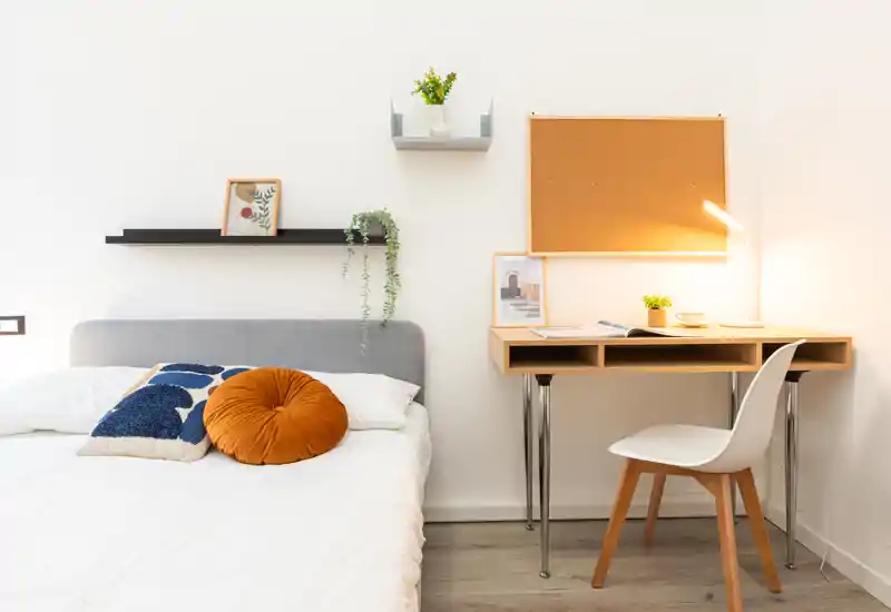 Camera da letto luminosa e moderna con letto ben rifatto, cuscini decorativi e un angolo studio compatto con scrivania in legno e lampada — ideale per comfort e lavoro da remoto.