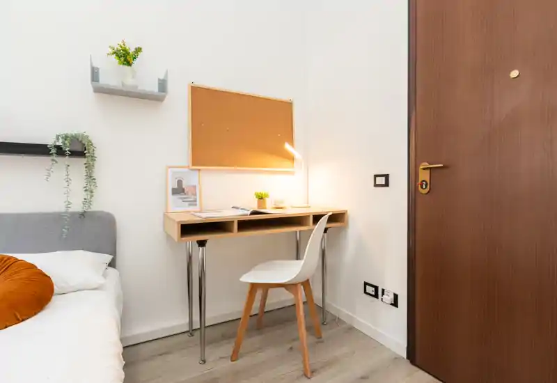 Bright, cozy bedroom corner with a single bed, bedside textiles, and a small desk with a chair and lamp — ideal for studying or working.