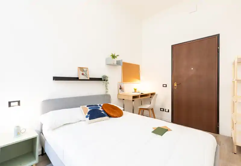 Camera da letto moderna e luminosa con un letto matrimoniale ben rifatto, cuscini decorativi, un piccolo comodino e una scrivania compatta vicino all'ingresso. La stanza è fresca e ben illuminata, perfetta per un soggiorno comodo.