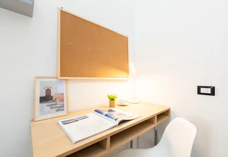 Bright, minimal desk corner with a light wood desk, corkboard, task lamp, small plant and an open book — a compact workspace ideal for studying or remote work.