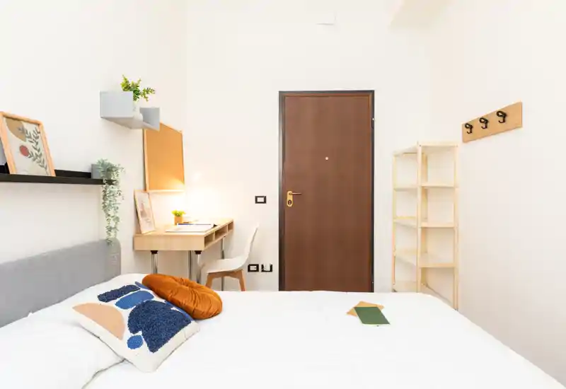 Camera da letto luminosa e ben arredata con letto in ordine, cuscini decorativi, una piccola scrivania con lampada e scaffali vicino alla porta d’ingresso — perfetta per un singolo o uno studente.