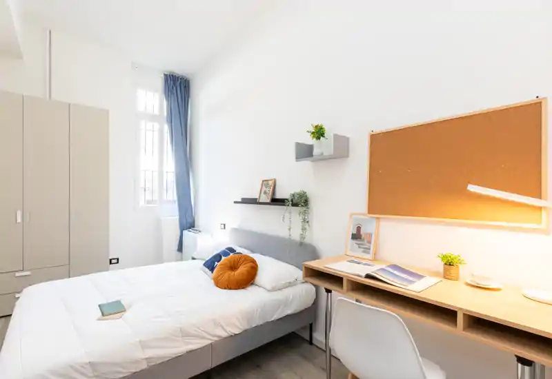 Bright, modern bedroom with a double bed, bedside shelving and a workspace with cork board — clean minimalist styling and plenty of natural light.