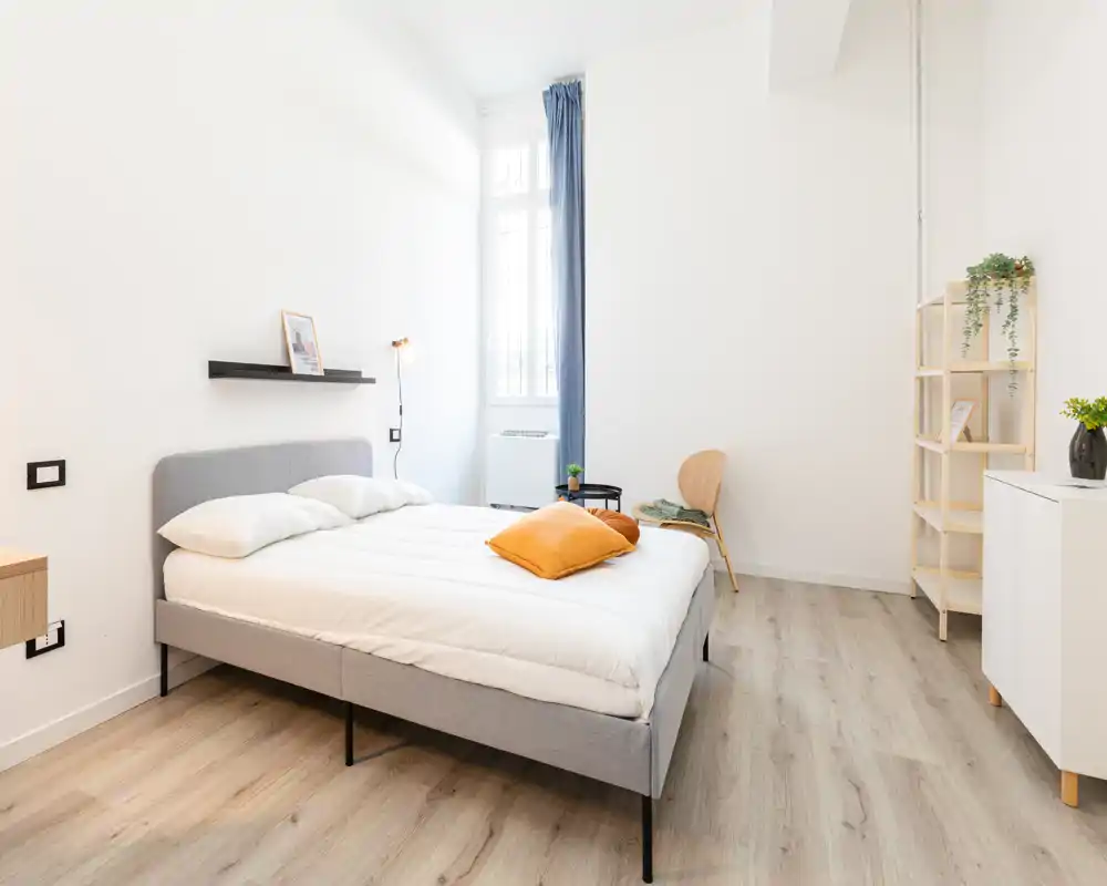Camera da letto moderna e luminosa con letto matrimoniale completo, pavimento in legno chiaro e arredo minimal. La grande finestra garantisce luce naturale e l'ambiente risulta arioso e accogliente.