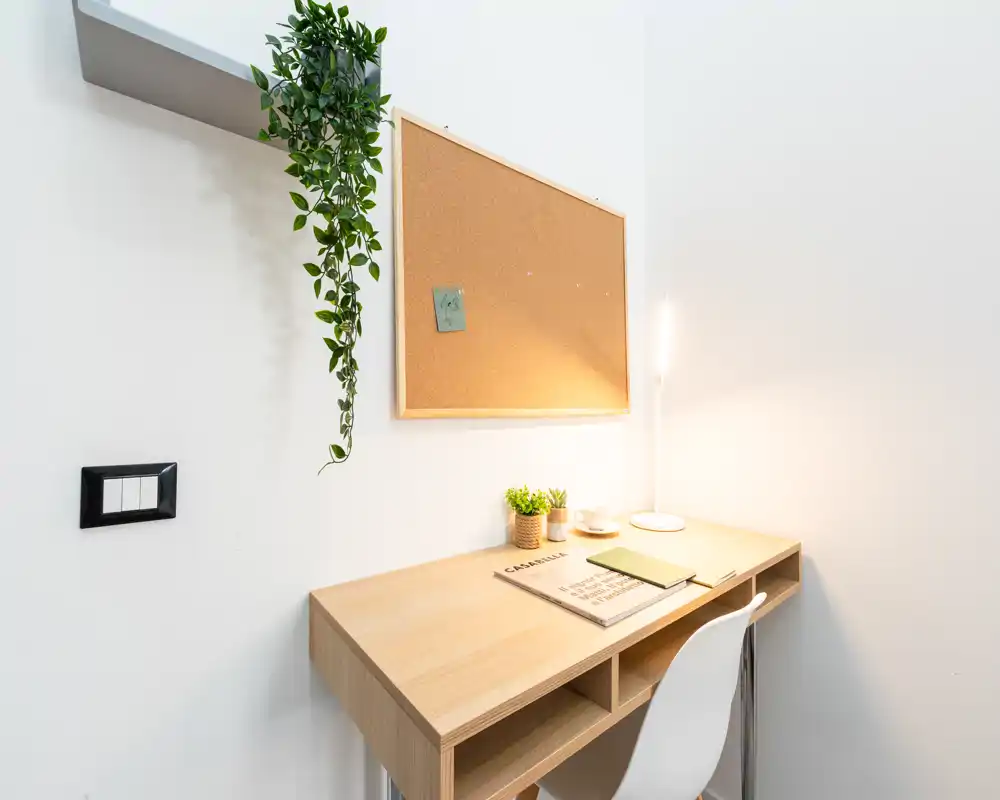 Angolo studio luminoso e minimale con scrivania in legno, lampada da tavolo, bacheca in sughero e piccole piante — ideale come postazione home office.
