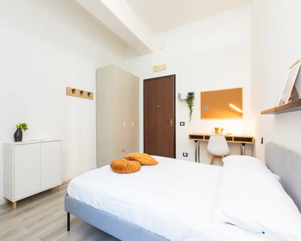 Camera da letto moderna e luminosa con letto matrimoniale ben rifatto, cuscini decorativi caldi e un piccolo angolo studio vicino alla porta. Linee pulite, pavimento in legno chiaro e arredamento minimale rendono l'ambiente accogliente e perfetto per affitti brevi.