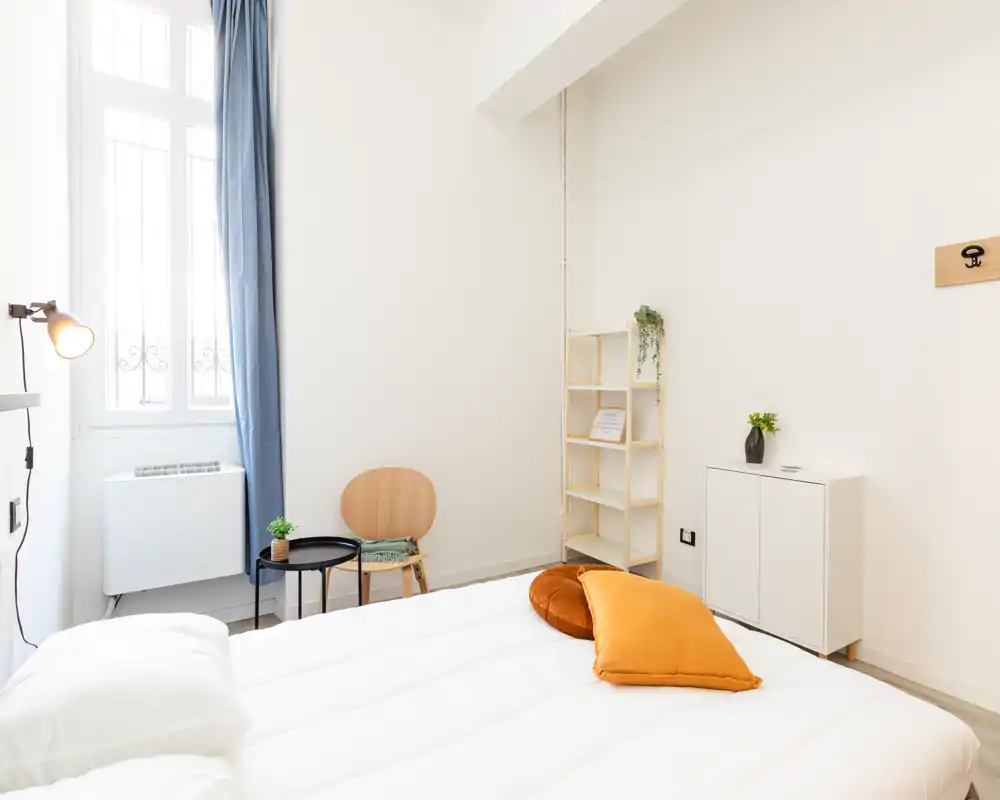 Camera da letto luminosa e minimale con letto ben rifatto, cuscini color senape, una piccola sedia in legno e una libreria a giorno — ideale per un soggiorno accogliente.