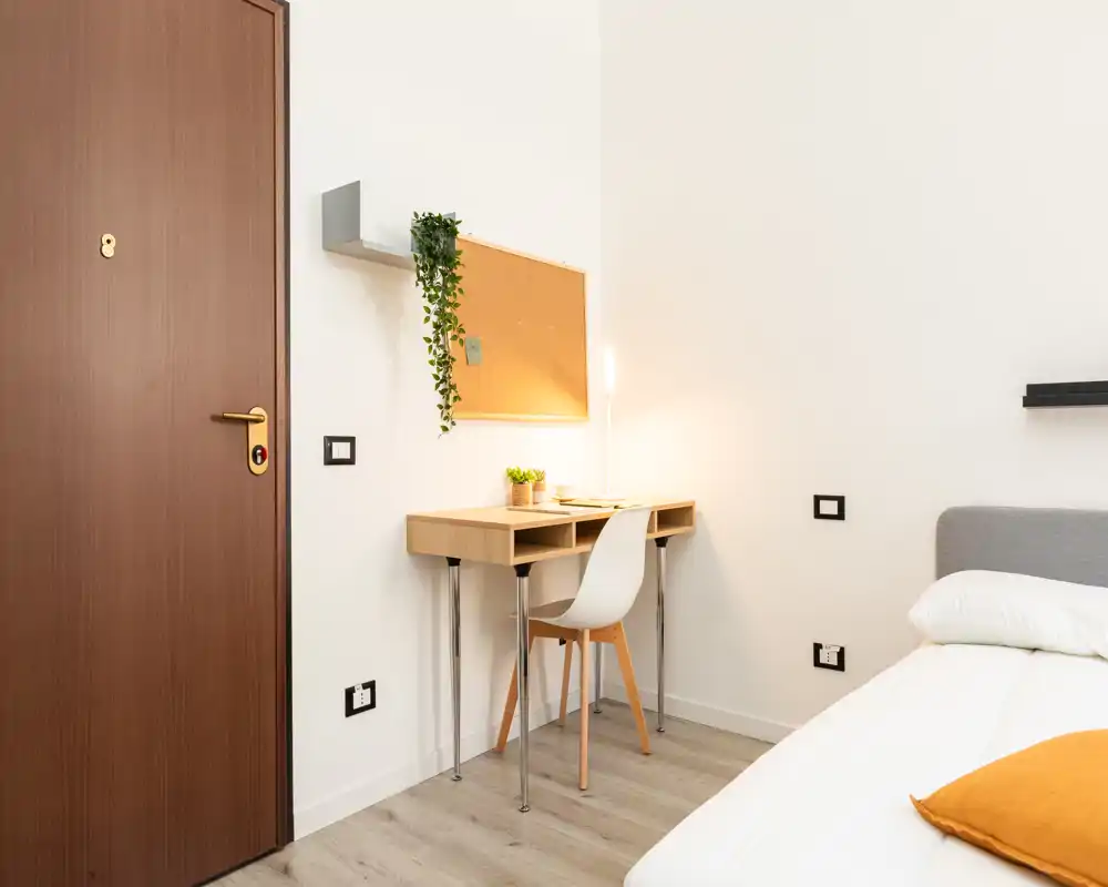 Angolo camera luminoso e minimal con letto singolo, piccolo scrittoio in legno e sedia vicino alla porta d'ingresso — design pulito, moderno e funzionale per affitti brevi.