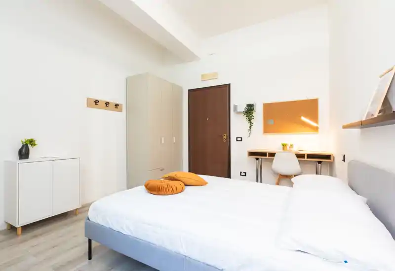 Camera da letto moderna e luminosa con letto matrimoniale ben rifatto, cuscini decorativi caldi e un piccolo angolo studio vicino alla porta. Linee pulite, pavimento in legno chiaro e arredamento minimale rendono l'ambiente accogliente e perfetto per affitti brevi.