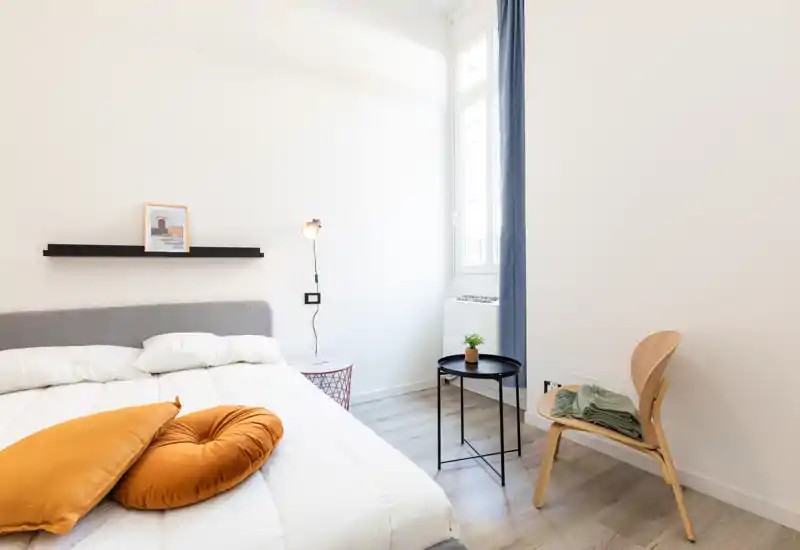 Camera da letto luminosa e moderna con un ampio letto in biancheria bianca, cuscini accoglienti e un piccolo tavolino vicino alla finestra — perfetta per annunci che puntano su uno stile minimal e confortevole.