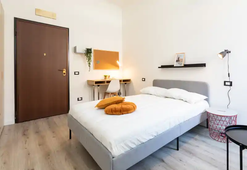 Camera da letto moderna e luminosa con letto matrimoniale ben rifatto, cuscini d'accento caldi e un piccolo angolo studio vicino alla porta — ideale per soggiorni brevi o lunghi.