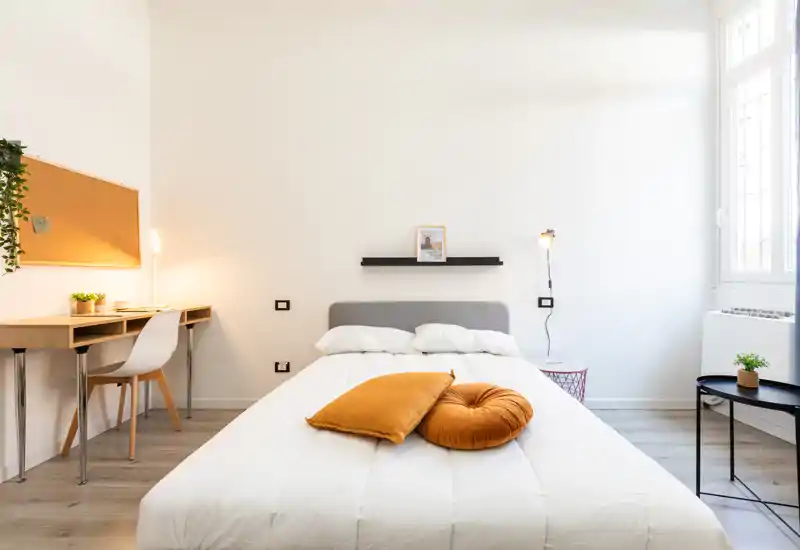 Camera da letto luminosa e minimal con letto matrimoniale al centro, cuscini dagli accenti caldi, una scrivania e una grande finestra che illumina l'ambiente.