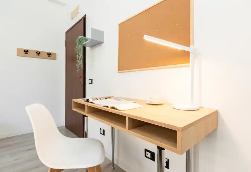 Small, bright entrance area with a slim wooden console used as a desk, a modern white chair, wall-mounted cork board and coat hooks; clean, minimalist styling and a desk lamp create a welcoming arrival space.