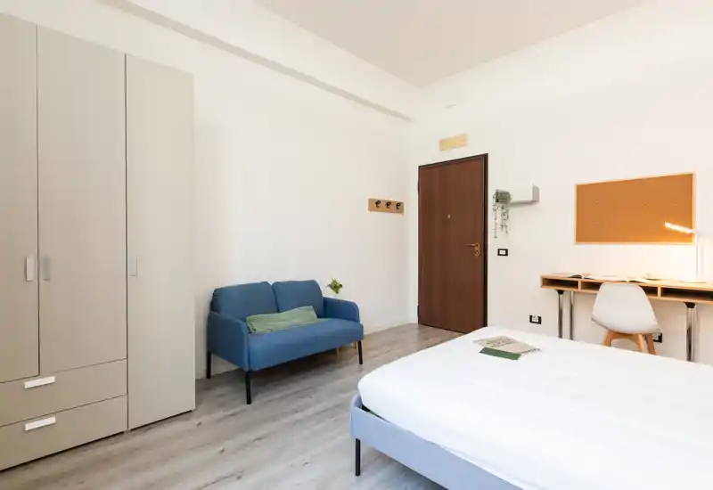 Bright, modern bedroom with a neatly made bed, a compact blue sofa, wardrobe and a small desk — ideal for a single renter or student.