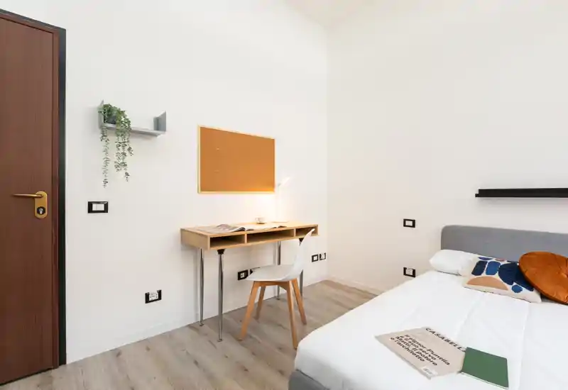 Bright, modern bedroom corner with a comfortable bed, simple desk and chair — ideal for a student or business traveler. The room features clean lines, light wood flooring and minimal, functional decor.