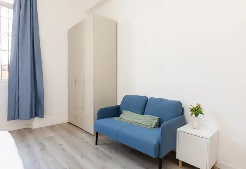 Small, bright living area with a compact blue two-seater sofa, wardrobe and a side table; clean, minimal styling and natural light from a window.