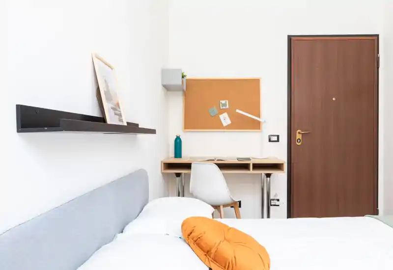 Bright, modern bedroom featuring a light grey upholstered headboard, white bedding with an orange accent cushion, and a small desk with chair by the door — ideal for a compact rental.