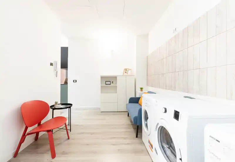 Bright, minimal living area with a small sofa, side table and seating; front-loading washing machines occupy the right side, creating a modern multipurpose space.