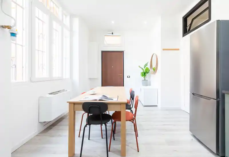 Bright, minimalist dining area with a wooden table and colorful chairs set beneath large windows; modern, airy styling ideal for listings.