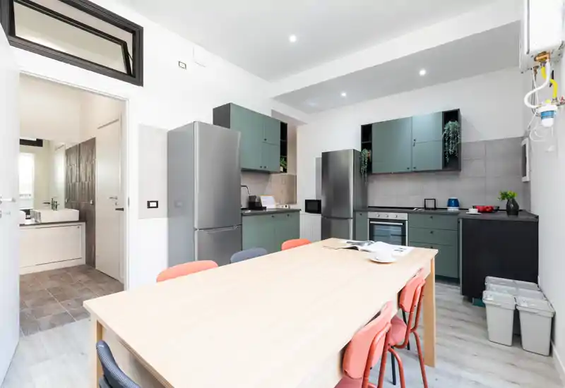 Modern open-plan kitchen with green cabinetry, two stainless refrigerators, built-in oven and a large wooden dining table with colorful chairs — bright, well-styled space ideal for shared meals.