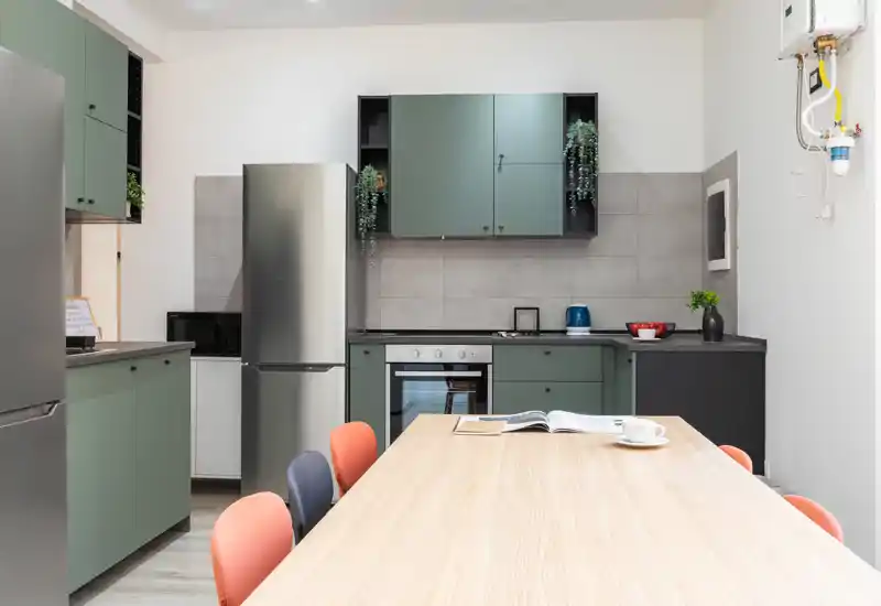 Modern kitchen with green cabinets, stainless appliances and a long wooden table with colorful chairs — bright, clean and ready for meals or remote work.