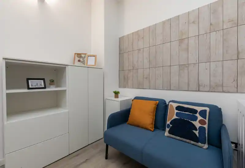 Compact, well-styled living area with a blue two-seater sofa, decorative cushions, wall panel art and clean white storage cabinets — bright and tidy space ideal for relaxing.