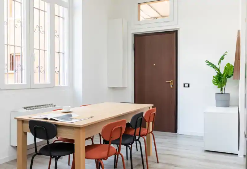Bright, minimal dining area with a wooden table and mixed-color chairs beside a large window; simple greenery and clean lines create a modern, welcoming space.