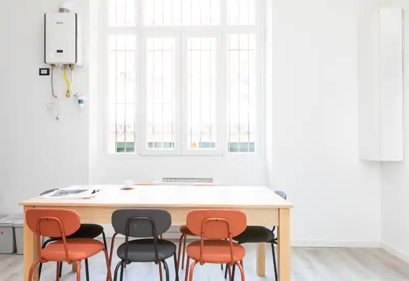 Bright, minimalist dining area with a light wood table and a mix of orange and dark chairs under a tall window, ideal for shared meals or workspace use.