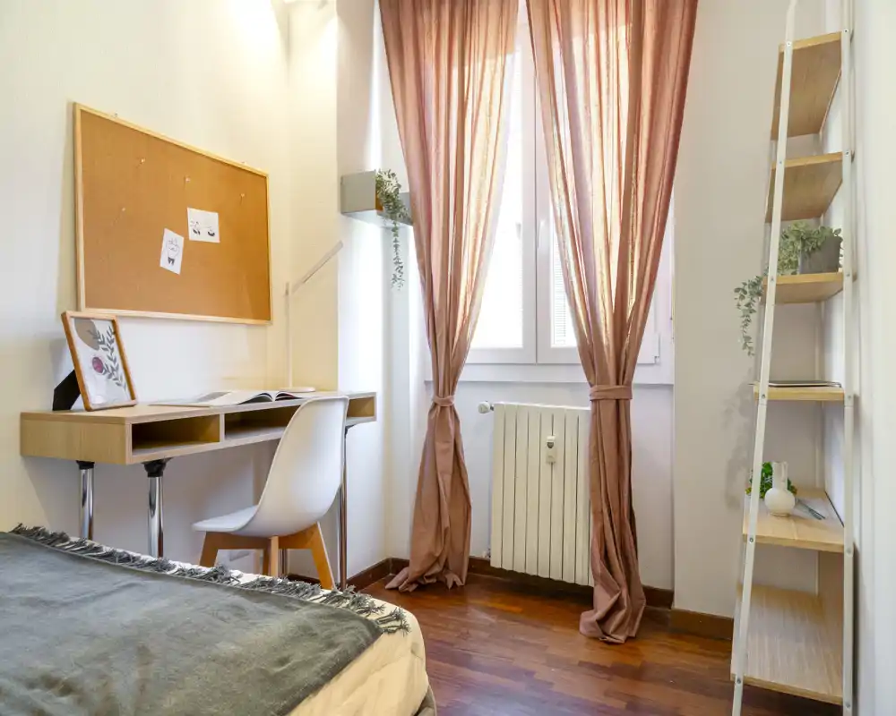 Rincón de dormitorio luminoso y bien decorado con cama visible, escritorio de madera con silla y ventana con cortinas rosa que difuminan la luz natural.