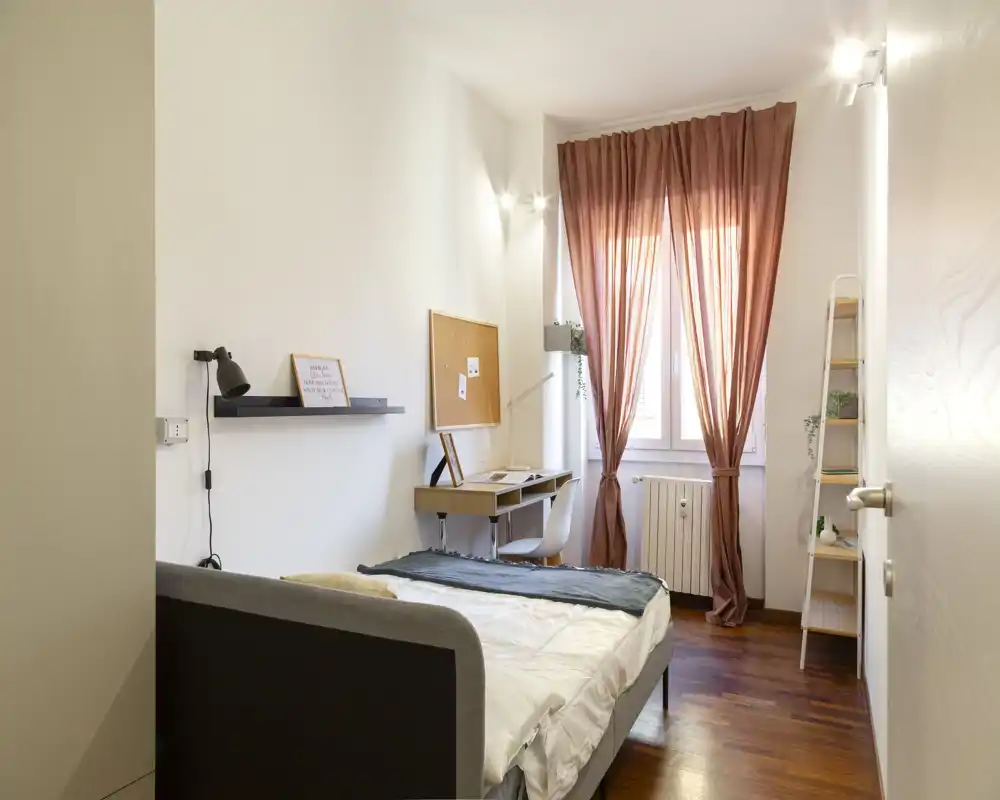 Dormitorio luminoso y ordenado con cama doble en primer plano, cortinas cálidas y un escritorio compacto junto a la ventana que crea un rincón acogedor y funcional.
