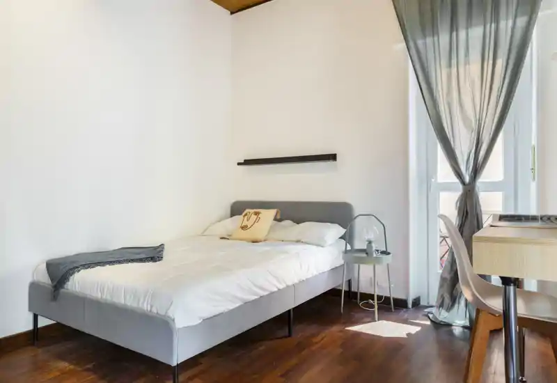 Bright, minimal bedroom with a modern grey bed, cozy linens and natural light from a large window leading to a balcony.