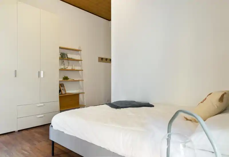 Minimal, well-lit bedroom with a comfortable double bed, simple wardrobe and shelving; neutral tones and wooden floor create a calm, modern atmosphere.