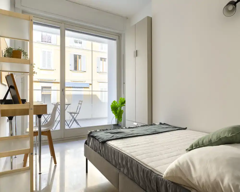 Bright, modern bedroom with a double bed in the foreground, large sliding glass doors leading to a balcony, a small desk and shelving unit — ideal for a comfortable city stay.