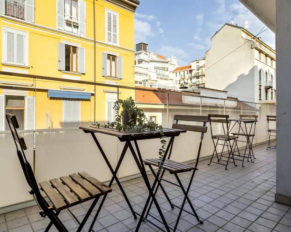 Sunny urban balcony with folding wooden tables and chairs, tiled floor and glass railing offering pleasant city views — ideal for morning coffee.