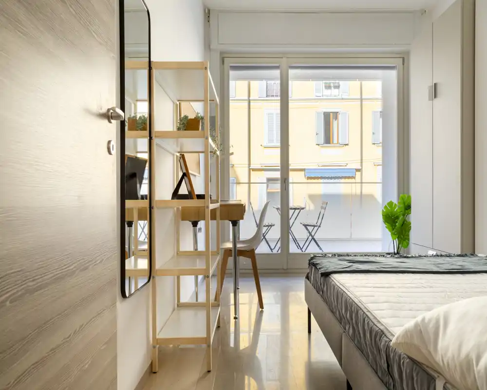 Bright, modern bedroom with a neatly made bed, a small work desk and shelves, and large sliding doors opening to a balcony with outdoor seating.