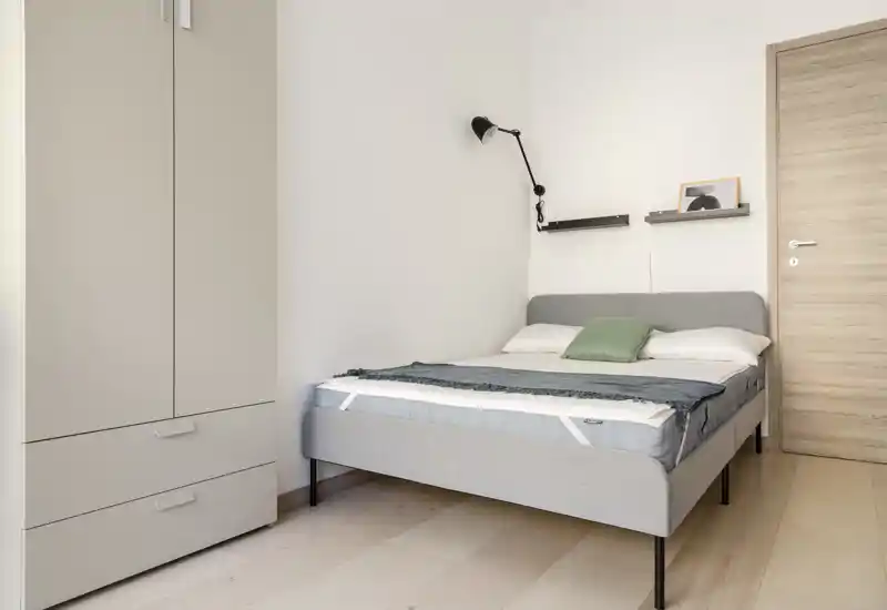Dormitorio luminoso y minimalista con cama doble bien hecha, lámpara de lectura en la pared y armario a la izquierda — estilo moderno y tranquilo, ideal para un anuncio de alquiler.