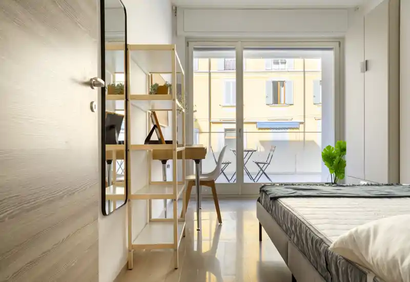 Dormitorio moderno y luminoso con la cama bien hecha, un pequeño escritorio con estanterías y grandes puertas correderas que dan a un balcón con sillas.