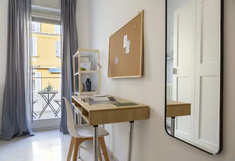 Small, bright bedroom corner with a wooden desk, chair and corkboard beside a tall window opening to a balcony.