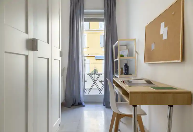 Bright, compact living area styled as a home office with a wooden desk, chair and shelving next to a tall window leading to a small balcony. Neutral tones, soft curtains and a cork noticeboard create a calm, functional workspace flooded with natural light.