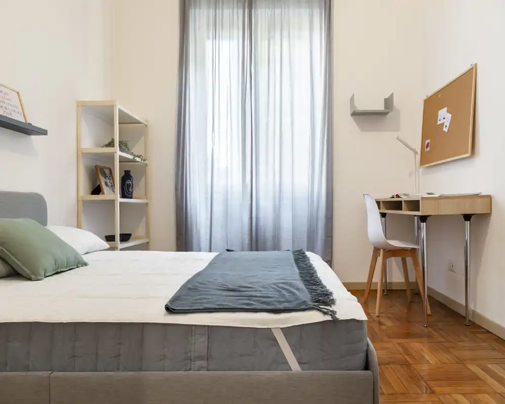 Dormitorio moderno y luminoso con cama doble bien hecha en primer plano, cortinas claras y un pequeño escritorio junto a la ventana — ideal para anuncios de alquiler.