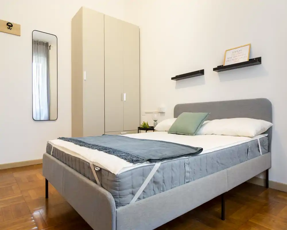 Dormitorio luminoso y ordenado con una cama tapizada gris moderna, almohadas suaves y una manta decorativa. Un armario, estantes en la pared y un espejo alto completan la distribución sencilla y acogedora.