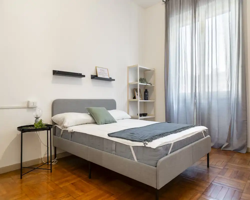 Dormitorio luminoso y minimalista con cama doble bien hecha, cabecero gris y luz natural que entra por una gran ventana con cortinas translúcidas.