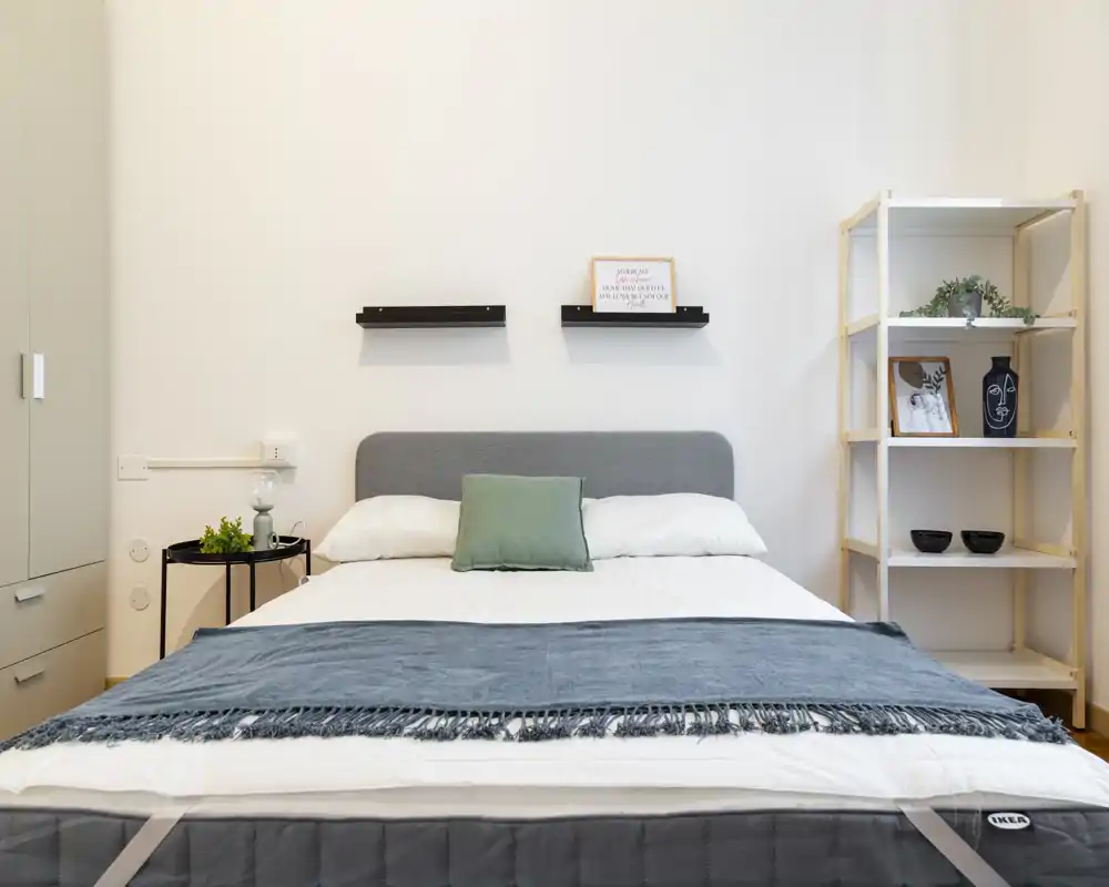 Dormitorio luminoso y minimalista con cama doble centrada, manta decorativa y cojín verde; estanterías sencillas y mesita completan el aspecto moderno y limpio.