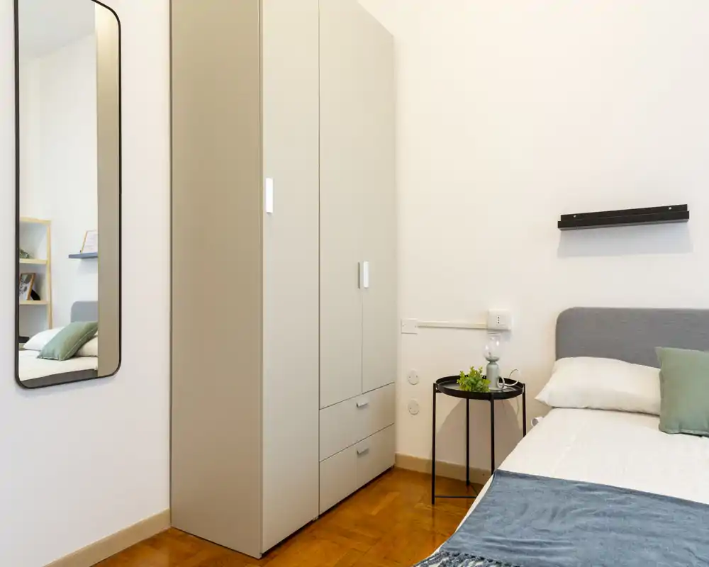 Dormitorio moderno y luminoso con cama bien hecha, cabecero gris, mesita y armario alto; estilo minimalista y suelo de madera cálido.