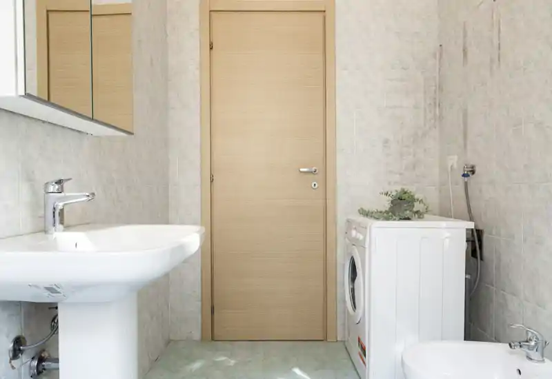 Salle de bains propre et compacte avec lavabo sur pied, bidet, machine à laver et murs carrelés neutres — lumière naturelle brillante et aménagement minimaliste fonctionnel.